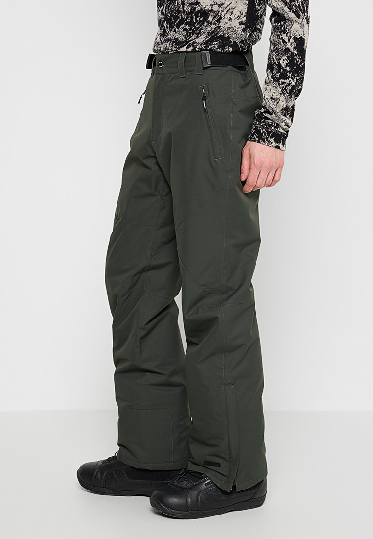 Grüne Schneehosen aus einem langlebigen Material. Sie verfügen über einen Gürtel, Reißverschlusstaschen und ein gerades Bein-Design. Getragen mit schwarzen Stiefeln.