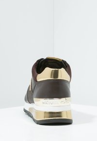 Sneaker marrone scuro con accenti dorati, intersuola bianca, suola nera e branding "MICHAEL KORS" sul tallone su uno sfondo bianco.