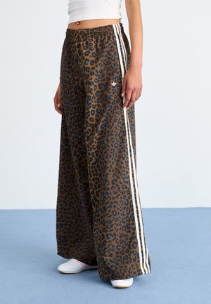 Osoba nosząca brązowe szerokie spodnie w panterkę z białymi paskami po bokach oraz białe trampki na niebieskiej podłodze.