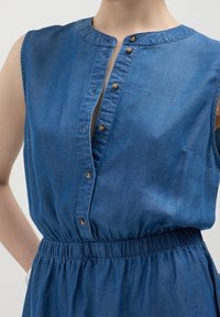 Sininen, hihaton fark dresses, jossa on pyöreä kaulus, napitettava etuosa ja joustava vyötärö, jota henkilö käyttää.