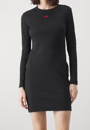 Kobieta ubrana w dopasowaną czarną mini sukienkę z długim rękawem, z małym czerwonym logo "HUGO" na klatce piersiowej.