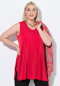 Top rosso senza maniche realizzato in tessuto leggero, con scollatura rotonda e vestibilità comoda. La modella tiene una sciarpa fantasia appoggiata sul braccio.
