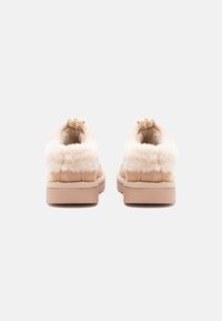 Beige slip-on shoes with a fluffy fabric upper, cushioned collar, and decorative gold accent on the back, featuring a tan rubber sole.