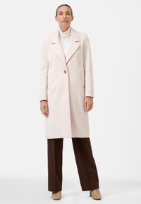 Lichtroze, gestructureerde jas met een ingesneden kraag, sluiting met één knoop en zijzakken, gecombineerd met brown broeken en beige schoenen.