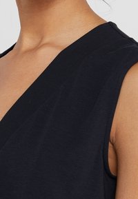 Haut sans manches noir en tissu doux, avec un décolleté en V profond et une texture lisse. Accent sur les détails des épaules et de l'encolure.