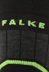 Calzino nero con texture a coste e accenti verdi. Presenta il marchio "FALKE" in evidenza vicino alla parte superiore e un bordo a righe verdi a contrasto.