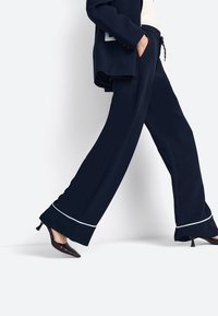 Tengerészkék bőszárú nadrág fehér szegéllyel a szegélyén, fényes bordó magas sarkú cipővel párosítva. Simogató anyag és lazított szabás.