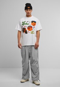 Hvit bomull T-skjorte med grafiske basketballdesign og tekst, kombinert med løse, lysegrå bukser og beige sneakers. Svart beanie-hatt.