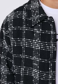 Zwarte textuurjack met wit geruite patroon, twee zwarte knopen, gedragen over een wit shirt.