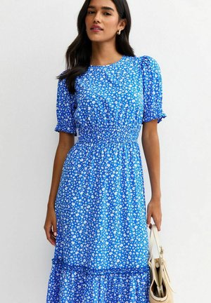 Mujer con un vestido midi azul con estampado floral blanco, mangas abullonadas, cintura entallada, sosteniendo un bolso beige sobre un fondo claro y liso.