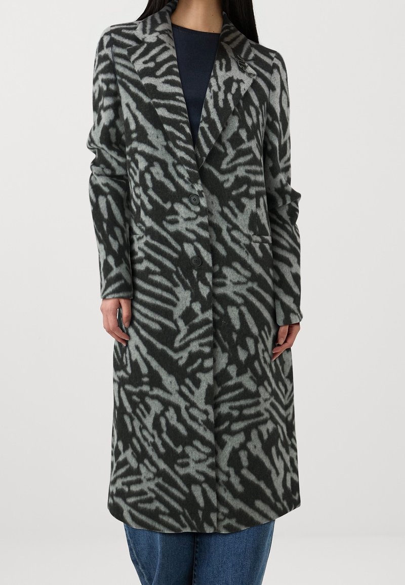 Long manteau noir et gris à motif avec de larges revers, deux boutons et des poches latérales, porté par-dessus un haut sombre et un jean bleu.