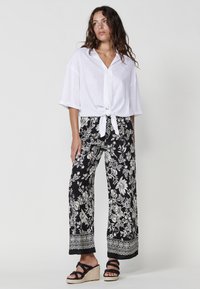 Camicia bianca a maniche corte, con bottoni e annodata in vita, abbinata a pantaloni larghi neri con motivo floreale e sandali con zeppa neri a strisce.