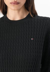 Pull noir avec un design en tricot torsadé texturé, présentant un col rond côtelé et un petit logo de marque en rouge, blanc et bleu sur la poitrine.