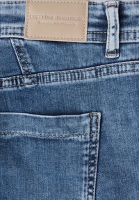 Tejido de denim azul con un acabado desgastado, que presenta un bolsillo parche y una etiqueta de cuero marrón cosida en la cinturilla.