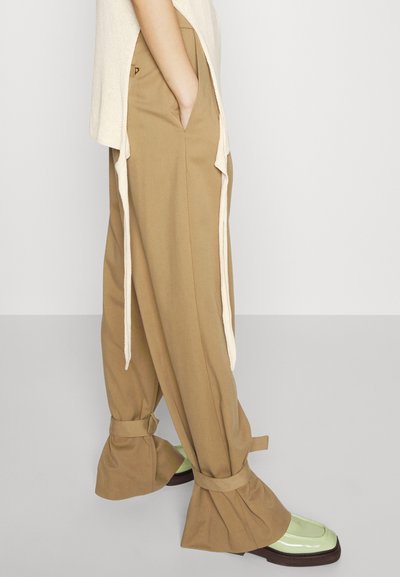 Beige színű, bő szárú nadrág, megkötött szegéllyel, oldalsó zsebekkel és kontrasztos elefántcsont színű részletekkel; zöld és barna lábbelivel párosítva.