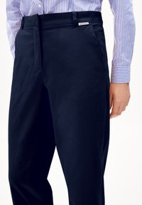 I pantaloni blu navy presentano un tessuto liscio e strutturato con una vestibilità slim, tasche laterali e un'etichetta sulla vita. Indossati con una camicia a righe.