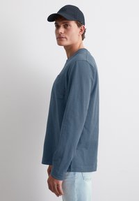 Langärmliges Hemd in Dunkelblau, aus strukturiertem Stoff, mit einer Vorderentasche und einer lässigen Passform. Kombiniert mit hellblauen Jeans und einer passenden Mütze.