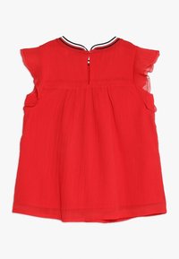 Haut sans manches rouge avec des épaules évasées, en tissu léger texturé, finition au dos avec une découpe en clé, et un col rayé contrasté.