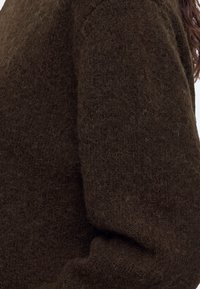Gros plan d’un tissu texturé duveteux marron foncé sur un vêtement couvrant le bras et l’épaule d’une personne.