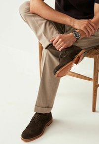 Braune Wildleder-Wingtip-Schuhe mit durchbrochenen Details, kombiniert mit hellbeigen Hosen. Der Schuh verfügt über eine strapazierfähige Gummisohle und ein Schnürdesign.