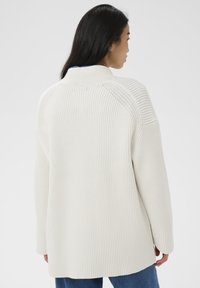 Žena s dlhými tmavými vlasmi, oblečená v bielom rebrovom úplete a modrých džínsoch, otočená chrbtom k fotoaparátu, na jednoduchom svetlom pozadí.