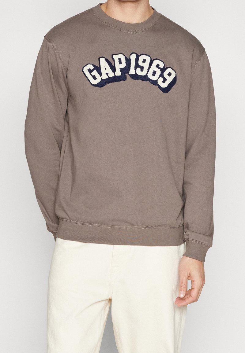 Sweatshirt marron avec "GAP 1969" brodé en bleu marine, col rond, manches longues, poignets et ourlet côtelés. Associé à un pantalon clair.