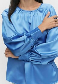 Satängblå blus med rynkad halsringning, långa ärmar och knappade manschetter; slät textur och avslappnad passform, idealisk för lagerpåläggning.