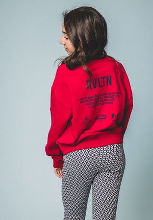 Rode sweatshirt met een ontspannen pasvorm, voorzien van marineblauwe tekst op de achterkant, gecombineerd met gestreepte, taps toelopende broek in een marineblauw en wit geometrisch ontwerp.