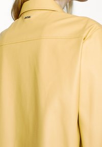 Femme portant une chemise jaune pâle à col avec un petit logo métallique BOSS dans le haut du dos, près de l'encolure.
