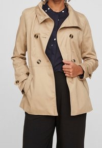 Beige trenchcoat met een kraag, dubbelzijdig ontwerp en donkere knopen. Heeft zijzakken en opgerolde mouwen over een marineblauwe blouse.