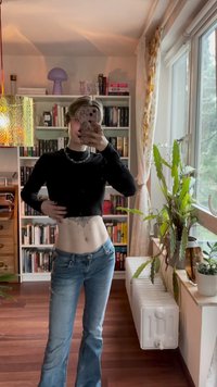 Suéter negro corto con cuello alto, combinado con jeans acampanados de un azul claro. Presenta un collar de cadena de plata y tatuajes visibles en el abdomen.