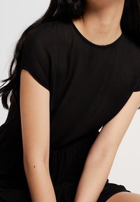 Schwarzes Kurzarmkleid mit lockerer Passform, rundem Halsausschnitt und gerafftem Taillenbereich, aus leichtem Stoff mit leicht strukturierter Oberfläche.