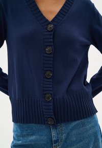 Marinblå cardigan med ribbat v-ringning och nederkant, med sex svarta knappar framtill och ribbade detaljer på ärmarna och midjan.