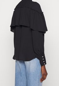 Schwarze Bluse mit gerafftem Overlay, langen Ärmeln und Knopfakzenten an den Bündchen. Leichter Stoff, lockere Passform und abgerundeter Saum.