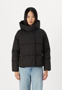 Zwarte pufferjack met een hoge hals en een vierkante pasvorm, met een gladde textuur en zichtbare naadlijnen, gedragen over lichte jeans.