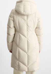 Vaalean beige-takkipuku, jossa on huppu, ja joka esittelee viistettyä tikkausta, pitkät hihat ja ribattuja hihansuita. Takana on tyylikäs, sileä pinta.