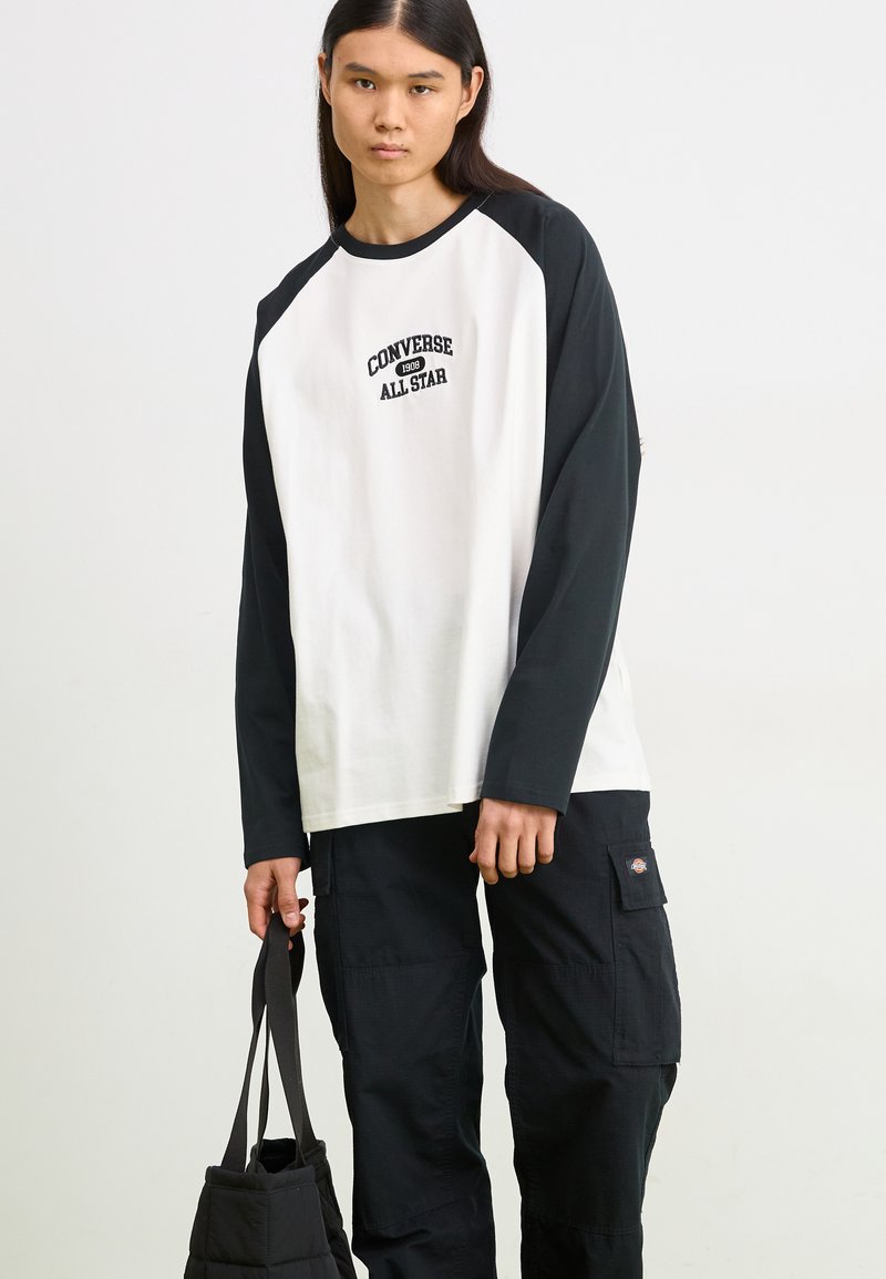 Chemise à manches longues blanche et noire avec des manches raglan, arborant le logo "Converse All Star" en noir sur la poitrine ; associée à un pantalon cargo noir.