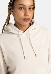 Beige hoodie made of soft material, featuring a drawstring hood, front pocket, and embossed branding on the chest. Silver aglets on drawstrings.