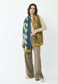 Wollmischung-Schal mit auffälligen blauen, grünen und orangefarbenen abstrakten Mustern und Fransen, über einem hellen Pullover und einer Hose drapiert.