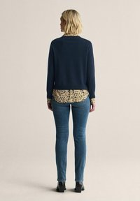 Marinblå tröja över en skjorta med gul blommönster, kombinerat med medelblå jeans och svarta anklar stövlar; släta texturer och lagerdesign.