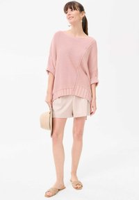 Roze oversized gebreide trui met een diagonaal naad en geribbelde zoom, gecombineerd met beige shorts en lichtbruine instapsandaaltjes. Eenvoudige handtas in de hand.