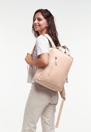 Sac à dos beige avec une surface texturée, poche avant zippée et deux bandoulières, porté par une personne vêtue d'une chemise gris clair.