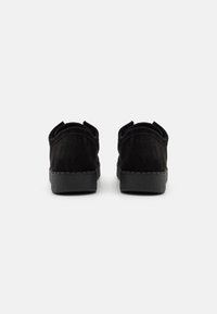 Chaussures en daim noir avec une semelle en caoutchouc texturée noire. Elles présentent des bords arrondis et deux languettes à l'arrière pour faciliter l'enfilage.