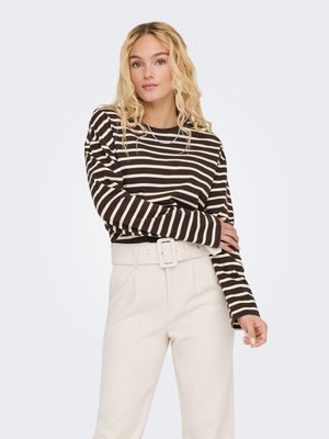 Mujer rubia con pelo ondulado que lleva una camiseta de manga larga a rayas en blanco y negro y pantalones beige de talle alto con un cinturón ancho.