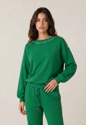 Junge Frau mit welligem dunklem Haar trägt einen grünen Sweatshirt und passende Hosen, die mit Perlendetails entlang der Kanten und Ärmel verziert sind.