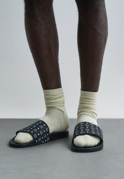 Black slip-on sandals with a patterned upper, worn over light-colored socks. Smooth texture, flat sole, and minimal design accents.