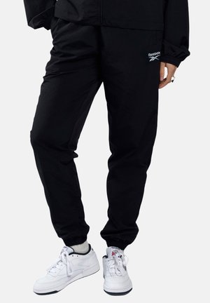 Persoon draagt zwarte Reebok trainingsbroek en witte Reebok sneakers met witte sokken tegen een effen achtergrond.