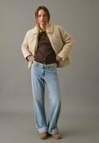 Mujer vistiendo una chaqueta acolchada beige sobre un suéter marrón, jeans de pierna ancha azul claro con puños y zapatos color canela, de pie contra un fondo gris.