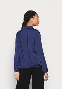 Camisa de mangas longas, azul-marinho, com uma superfície texturizada, colarinho e bainha elástica. Apresenta punhos com botão e um corte solto.