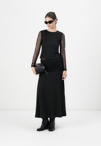 Black long-sleeve top with sheer mesh sleeves, paired with a black ruched maxi skirt and black ankle boots; carrying a small textured handbag.